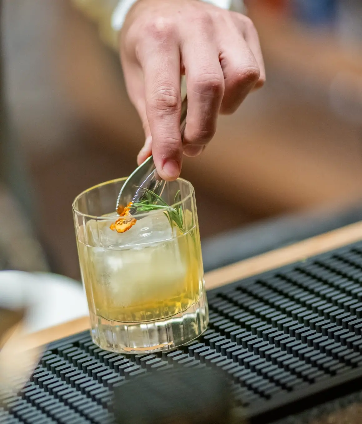 Mano che rifinisce un cocktail con rosmarino e scorza d’agrume, servito in bicchiere basso con ghiaccio.