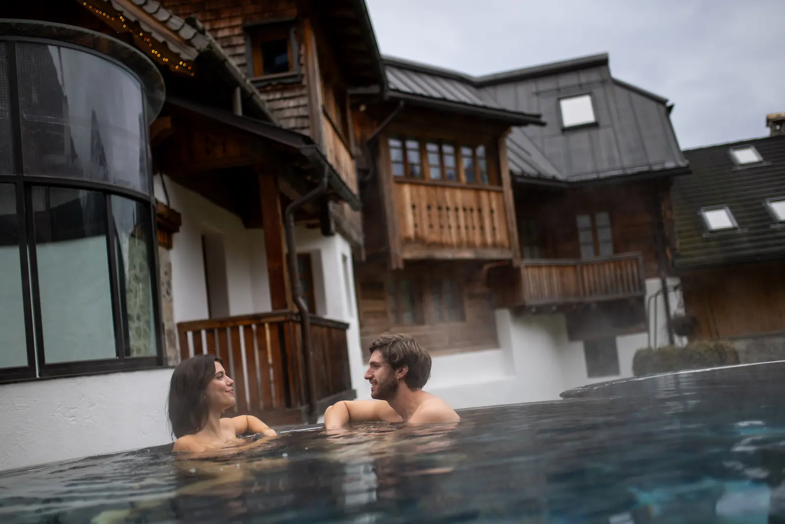 Coppia che si rilassa in una piscina esterna riscaldata, circondata da chalet alpini in legno.