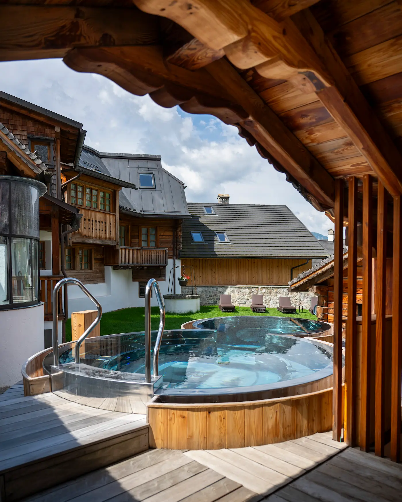 Piscina esterna circolare in legno con idromassaggio, immersa tra chalet alpini e relax all’aperto.