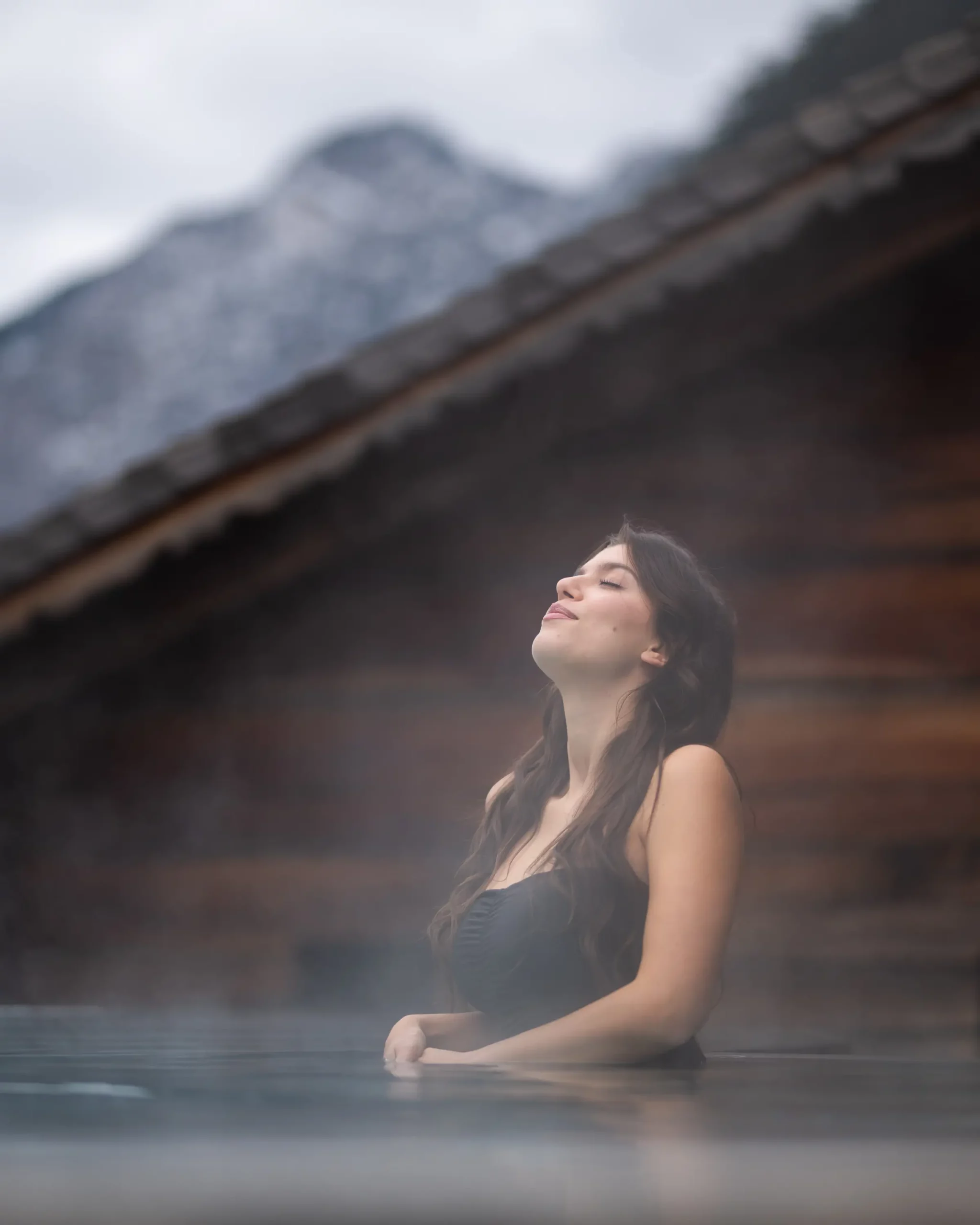 Donna che si rilassa in una piscina esterna riscaldata, avvolta dal vapore con sfondo alpino innevato.